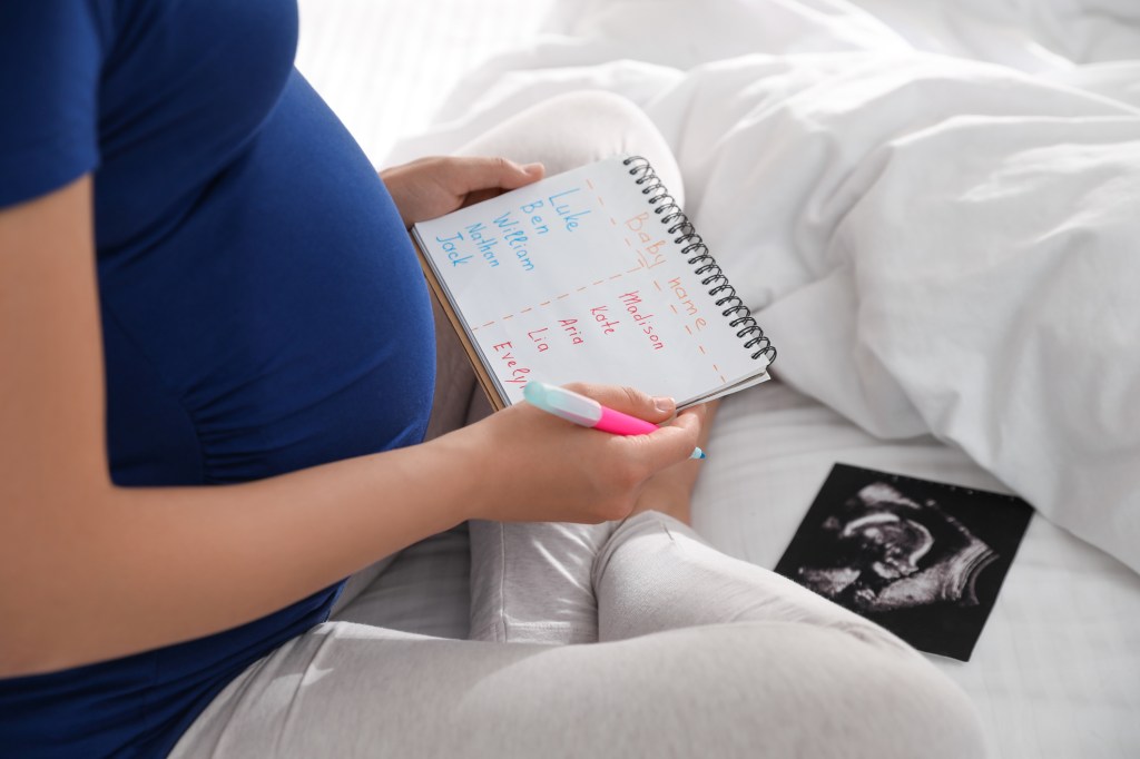 Pregnant woman holding a list of baby names and sonogram, sitting on a bed